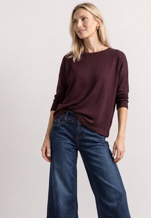 Lyshåret kvinde iført en mørk burgunderfarvet langærmet sweater og løstsiddende blå denimjeans, stående mod en ensfarvet lys baggrund.