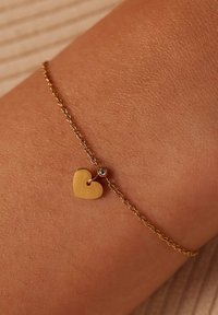 Gouden kettingarmband met een kleine hartvormige bedel en een heldere rhinestone accent. De ketting heeft een delicate, gestructureerde schakels ontwerp.