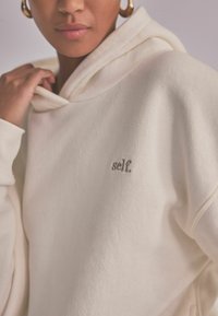 Gai beidža kapuce, izgatavota no mīksta materiāla, ar priekšējo kabatu un mazām izšūtām "self." logo pelēkajā krāsā. Dizains ietver kapuci ar auklu.