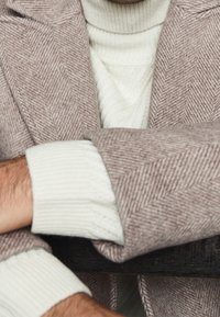 Un cappotto a motivo chevron marrone si adagia su un maglione bianco lavorato a maglia, con polsini a coste visibili sulle maniche.