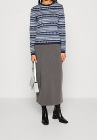 Pull en tricot avec un motif géométrique bleu et blanc, associé à une longue jupe grise. Sac à main argenté et bottines à talons carrés noires.