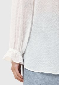 Hvid tekstureret bluse med lange, løse ærmer og flæseærmer. Stoffet har et subtilt ternet mønster og er let.