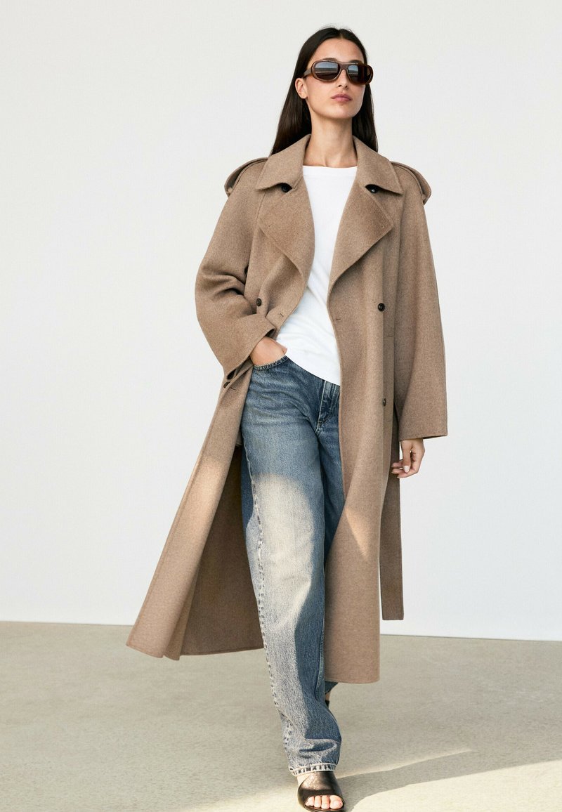 Woman wearing long camel coat over white shirt and blue jeans, with black sunglasses and black open-toe sandals, walking indoors on light floor.