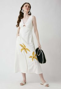 Weißes Kleid mit V-Ausschnitt, Frontausschnitt und gelb gestickten Palmblättern. Das Modell hält eine schwarze Tasche und trägt flache beige Sandalen.