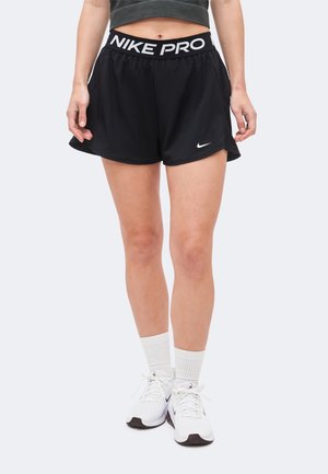 Kobieta w czarnych sportowych szortach Nike Pro, białych butach sportowych Nike i białych skarpetkach, stoi na tle jednolitego jasnoszarego tła.