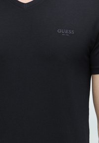 T-shirt noir col V avec le logo "GUESS EST. 1981" brodé sur le côté gauche de la poitrine, porté par une personne.