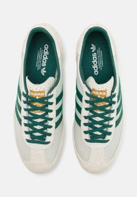 Vita lädersneakers med gröna detaljer, tre ränder, texturerad tåhätta och en mjuk, fluffig sula. Gyllene logotyp nära skosnören.