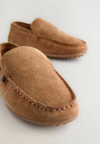 Mocassins en suede marron avec une texture lisse, bout arrondi, couture décorative et un petit bouton sur le côté. Semelle en caoutchouc flexible.