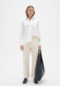 Camisa blanca de botones, pantalones de pierna ancha crema y un gran bolso de cuero negro; el modelo lleva zapatos negros, fondo neutro.