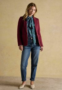 Naiste burgundi värvi bleiser, sinise mustriga pluus koos lipsuga, kõrge vöökohtadega teksad ja beežid madala kontsaga kingad seisavad beežil taustal.