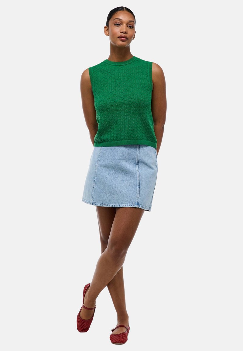 Haut sans manches en tricot vert associé à une mini-jupe en denim clair et des chaussures plates texturées rouges. Fond uni.
