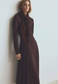 Robe longue marron avec manches longues et col montant, agrémentée d'une écharpe nouée à l'épaule. Texture lisse, design fluide.