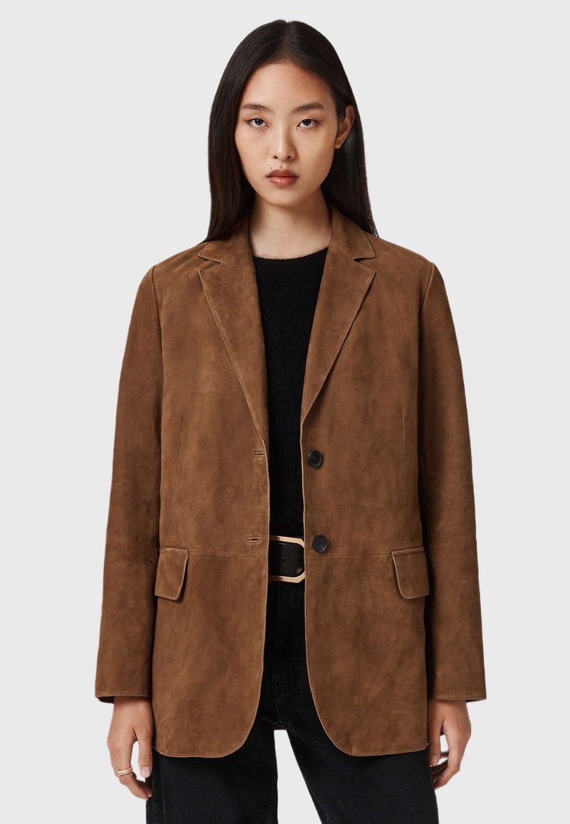 Bruine suède blazer met gelapte revers, twee knopen en zijzakken. Draag over een zwarte trui en zwarte jeans. Soepele textuur.