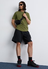 Un jeune homme se tient contre un mur uni, portant un t-shirt vert olive, un short noir, des lunettes de soleil et un sac bandoulière noir.