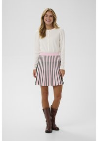 Fată tânără zâmbitoare, îmbrăcată cu o bluză albă cu nasturi și mâneci lungi, o fustă roz cu dungi verticale negre și cizme maro până la jumătatea gambei, pe un fundal simplu.