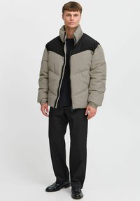 Pufferjack in lichtgrijs met zwarte accenten, voorzien van een hoge kraag, ritssluiting en gequilt ontwerp. Gecombineerd met zwarte broeken en schoenen.