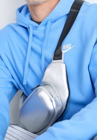 Stříbrná polostrukturovaná kabelka přes rameno s lesklým povrchem, která má černý popruh a zipový detail, opírá se o modrou mikinu s kapucí.