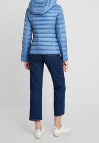 Niebieska puchowa kurtka z poziomym pikowaniem, z kapturem, o gładkiej, błyszczącej fakturze. Noszona z ciemnymi dżinsami i białymi trampkami.