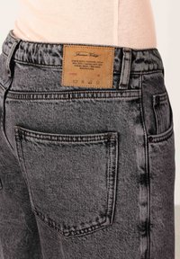 Tuva melnu skābās mazgāšanas džinsu aizmugures kabata ar ādas zīmola etiķeti un šuvju detaļām uz personas, kas valkā gaiši rozā virskreklu.