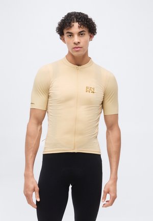 Άνδρας με σγουρά μαλλιά που φοράει μπεζ ποδηλατική φανέλα με κοντά μανίκια και μαύρο στενό παντελόνι, στέκεται μπροστά σε απλό λευκό φόντο.