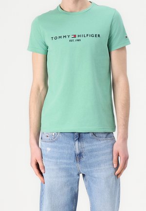 Osoba oblečená v mátově zeleném tričku Tommy Hilfiger s logem a světle modrých džínách na jednobarevném bílém pozadí.