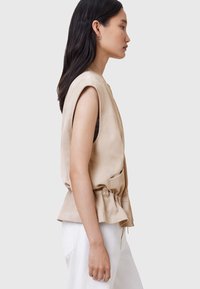 Gilet senza maniche beige in pelle, con una vestibilità rilassata, vita cinte e tasche laterali. Indossato sopra pantaloni bianchi.