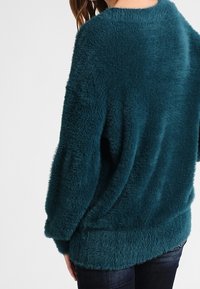 Turkos fluffig tröja med avslappnad passform, rund hals och ribbad nederkant. Texturen ser mjuk och plyschig ut. Lämplig för avslappnad användning.