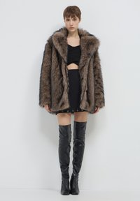 Donna che indossa un grande cappotto di pelliccia marrone, un crop top nero, una mini gonna nera con occhielli e stivali in pelle neri fino alla coscia, in piedi contro un muro bianco.