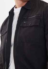 Veste noire à fermeture éclair avec deux poches avant, une poche poitrine zippée et un tissu texturé. Comprend un col et une chemise intérieure blanche.