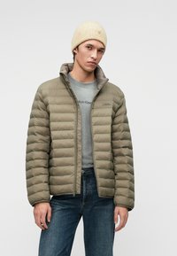 Žalia alyvuogių spalvos pūkinė striukė su horizontaliomis siūlėmis, su užtrauktuku ir aukštu apykakle, dėvima ant pilkos marškinėlių ir mėlynų džinsų, papildyta kreminės spalvos kepure.