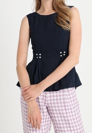 Top peplum sans manches bleu marine avec des accents de perles à la taille, associé à un pantalon à motifs pied-de-poule rose clair et blanc.