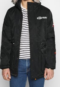 Parka negra impermeable con capucha forrada de piel sintética desmontable, botones a presión y dos bolsillos frontales. Presenta una capa interior a rayas y un diseño de logotipo.
