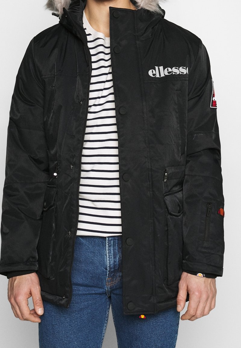 Parka negra impermeable con capucha forrada de piel sintética desmontable, botones a presión y dos bolsillos frontales. Presenta una capa interior a rayas y un diseño de logotipo.