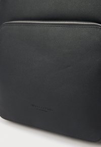 Svart lær ryggsekk med teksturert overflate, langstrakt form og en glidelås foran. Diskret merkevare plassering på forsiden.