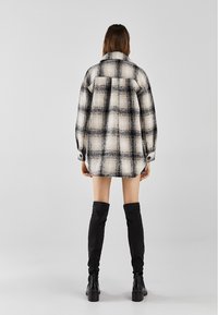 Nadměrná černobílá kostkovaná košile s límečkem, manžetami na knoflíky a texturovanou tkaninou, nošená přes černé kozačky sahající nad kolena.