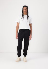 Svarte joggebukser med elastisk midje, kombinert med en hvit T-skjorte med korte ermer. Antrekket er fullført med kremfargede joggesko.