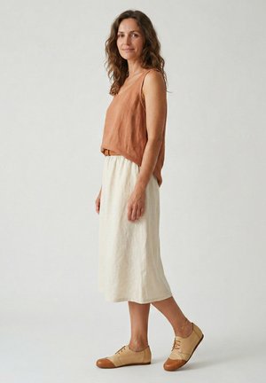 Vrouw staand zijwaarts, gekleed in een mouwloze bruine top, witte midirok en beige oxford schoenen tegen een effen achtergrond.