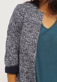 Námořnická a bílá texturovaná bunda s kulatým výstřihem a vyhrnutými rukávy, nošená přes teal top s výstřihem do V. Žádné viditelné zapínání.