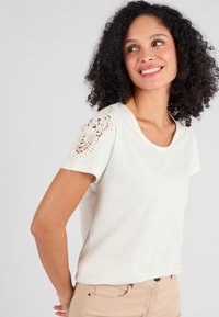 Top blanco de manga corta con escote redondo, que presenta un intrincado detalle de encaje en el hombro. Tela suave y ligera con una textura suave.
