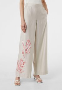 Beżowe spodnie o szerokich nogawkach z elastycznym pasem, zdobione czerwoną zieloną motywem po lewej stronie. Noszone z białymi sandałami na paskach.