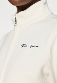 Sudadera blanca con cremallera y cuello elevado, que presenta un logo negro de "Champion" en el área izquierda del pecho, confeccionada con una tela texturizada.