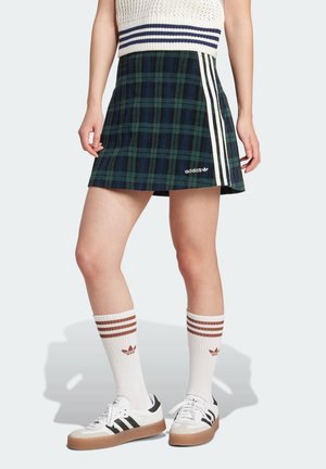 Geruite rok in donkergroen en marineblauw met witte zijkanten en een Adidas-logo, gecombineerd met witte sokken met bruine strepen en zwarte sneakers.