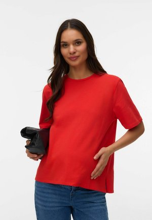 Kvinde med langt mørkt hår iført en rød kortærmet skjorte og blå jeans, der holder en sammenfoldet sort læderclutch i den ene hånd.