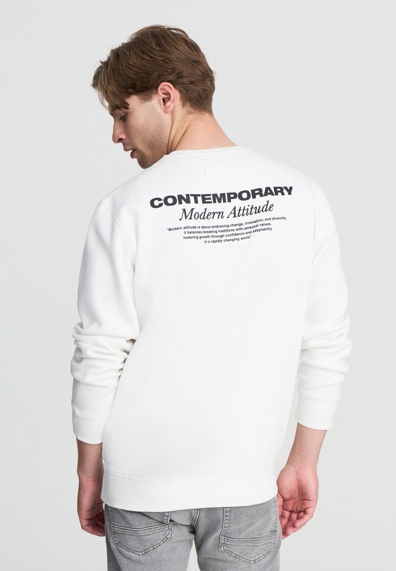 Felpa bianca con testo stampato in nero sul retro. Maniche e orlo a costine, vestibilità rilassata. Accoppiata con jeans grigi.