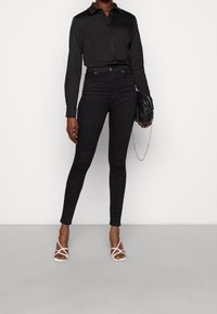 Schwarzes Hemd mit Knöpfen, hoch taillierte schwarze Skinny-Jeans und klare Riemchen-Absätze. Accessoires umfassen eine kleine schwarze Handtasche mit Kette.