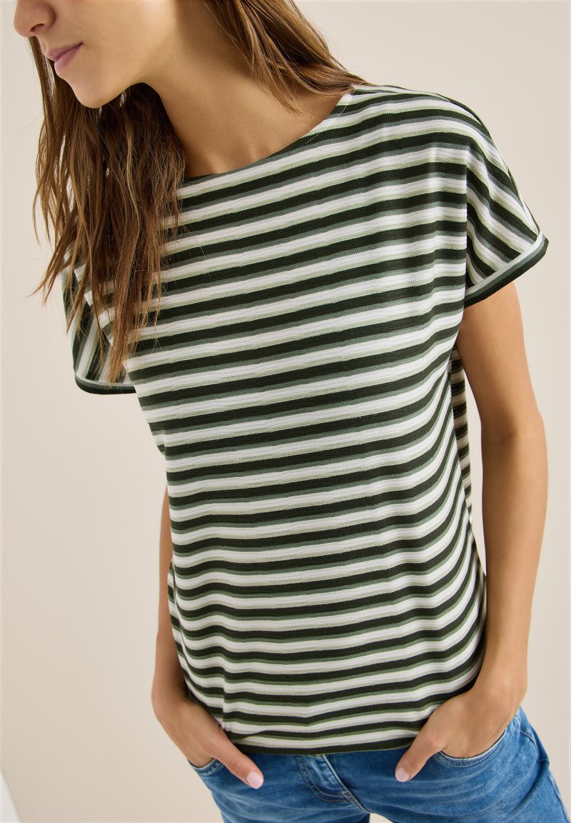 Femme portant un t-shirt à manches courtes à rayures horizontales vertes et blanches, les mains dans les poches d'un jean bleu, légèrement tournée vers la gauche.