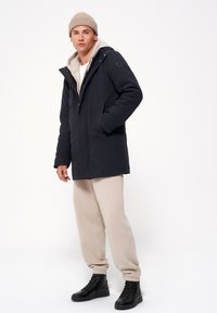Granatowa parka z kapturem, przednimi kieszeniami i beżowym wnętrzem, w połączeniu z beżowymi spodniami dresowymi i czarnymi botkami. Ciepły i codzienny design.