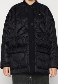 Black quilted jacket featuring a button-up front, round collar, and two front pockets. Made of shiny fabric with a smooth texture.