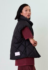 Schwarze gesteppte Puffer-Weste mit hohem Kragen, seitlichen Schlitzen und einem weißen Etikett. Getragen über einem kastanienbraunen kurzärmeligen Hemd, das eine glatte Textur aufweist.
