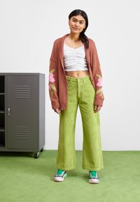 Cárdigan marrón de punto con diseño floral, top corto arrugado blanco y pantalones de pana verde de pierna ancha. Zapatillas verdes y rosas completan el look.
